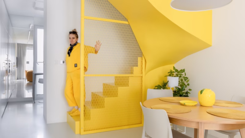 Yellow interior with metal mesh by the stairs, round wooden table with plants.