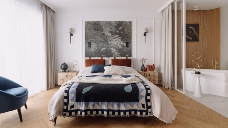 Bed with a geometric blanket and rust-coloured cushions in front of a large painting.