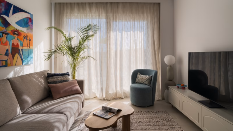 Living room with a beige sofa and a tall plant by sheer curtains.