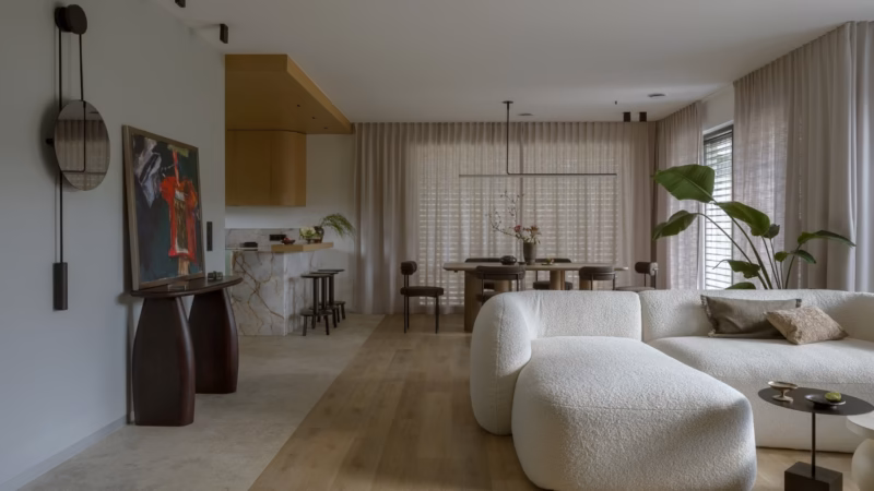 A living room with a curved, light-coloured sofa, a dining area and a wooden fitted kitchen