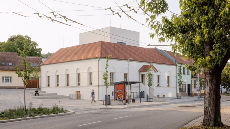 A white building with a red roof and a bus stop with a person sitting there